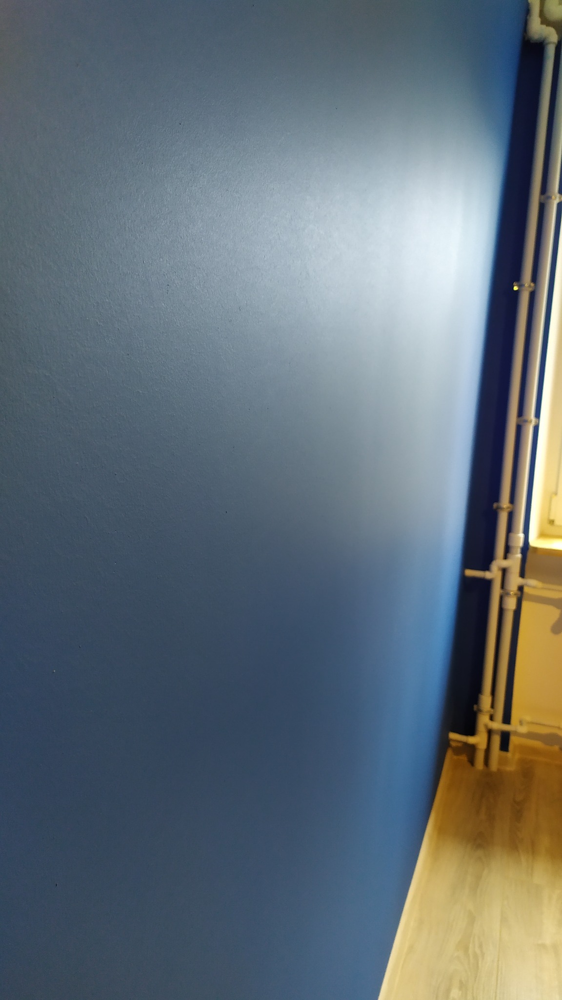 Świeżo pomalowana, jednolita, błękitna ściana z widocznymi rurami grzewczymi przy krawędzi i fragmentem drewnianej podłogi.