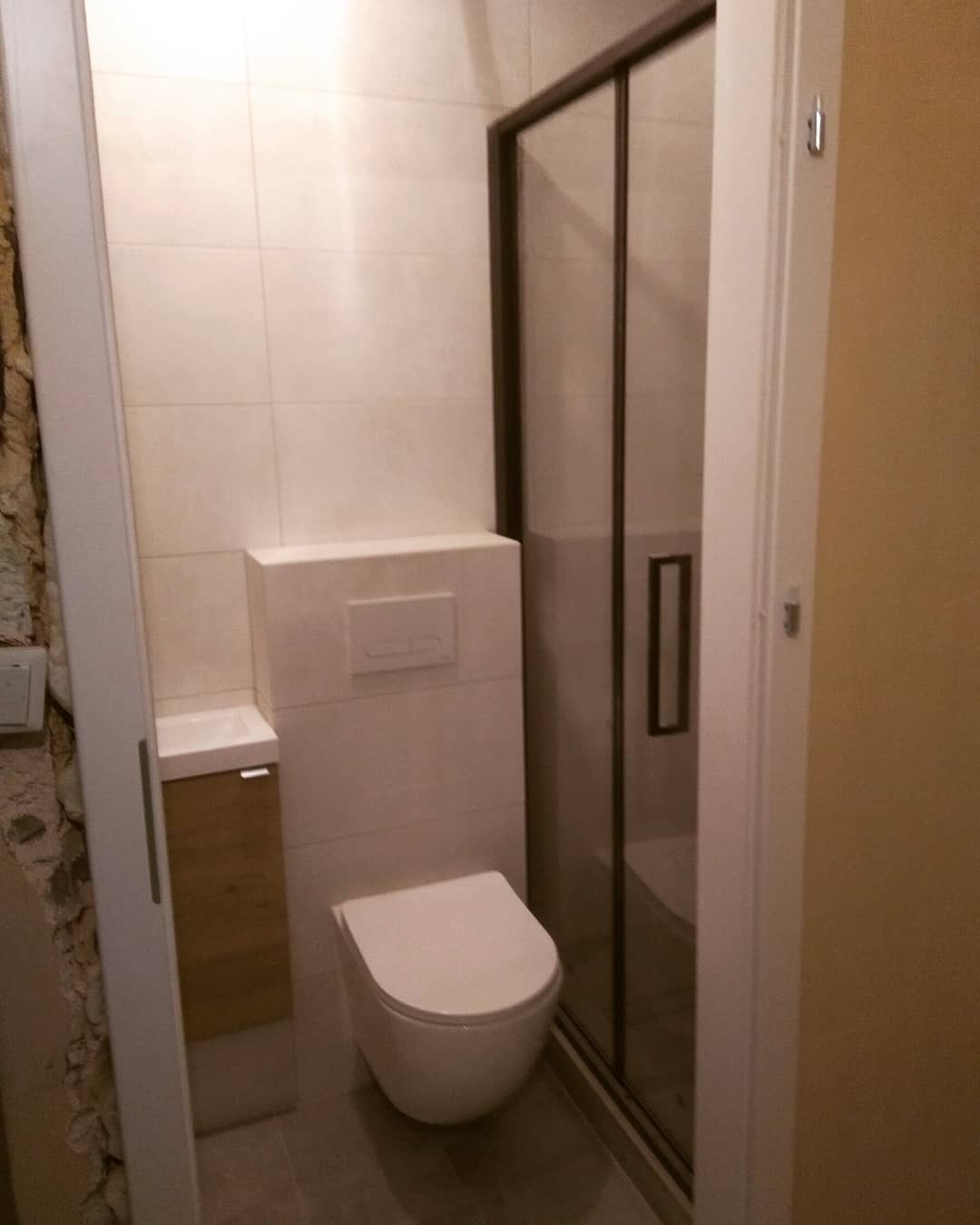 Nowoczesna łazienka z beżowymi płytkami, kabiną prysznicową z ciemną ramą, wiszącą toaletą i szafką z umywalką z drewnianym frontem.