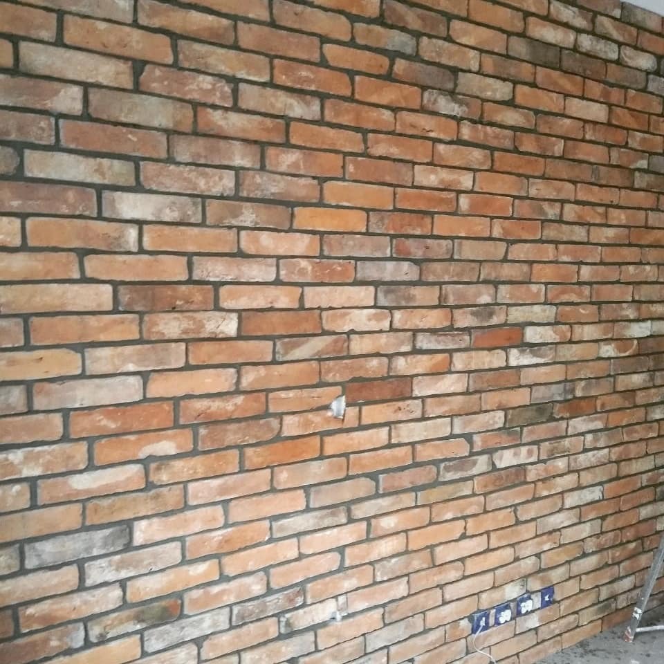 Ściana z cegieł klinkierowych w odcieniach czerwieni i brązu, z widocznymi fugami w kolorze grafitowym, gniazdka elektryczne przy podłodze, fragment drabiny w prawym dolnym rogu.