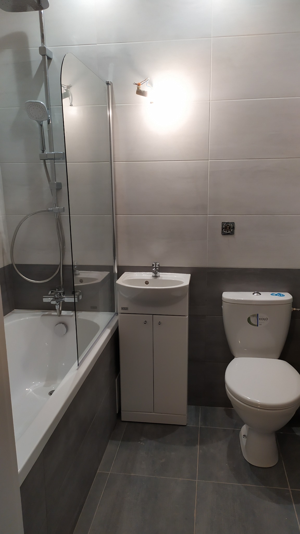 Nowoczesna łazienka z wanną z parawanem, umywalką z szafką i toaletą, wykończona szarymi i białymi płytkami, oświetlona punktowym światłem.