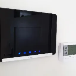 Panel sterowania Vidos z podświetlanymi ikonami i termostat De Dietrich zamontowane na białej ścianie, widoczne odbicie drzwi w ekranie.