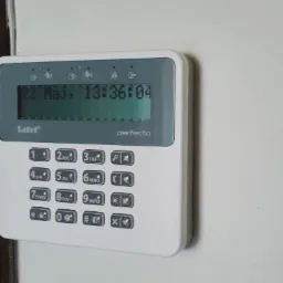 Panel sterowania alarmem SateLite Perfecta z wyświetlaczem pokazującym datę i godzinę, zamontowany na jasnej ścianie obok ciemnego elementu drzwi.