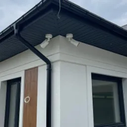 Dwie białe kamery monitoringu zamontowane pod okapem budynku z białą elewacją, widoczne kable oraz rynna.