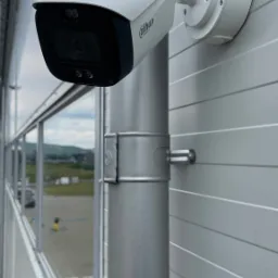 Zewnętrzna kamera monitoringu Dahua zamontowana na metalowym słupku przy ścianie budynku z widokiem na parking i odległe wzgórza.