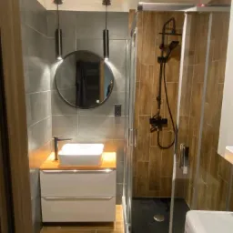 Nowoczesna łazienka z szarymi płytkami, okrągłym lustrem podświetlanym lampami, białą umywalką nablatową na drewnianym blacie i kabiną prysznicową z czarną armaturą.