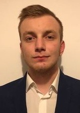 Portret młodego mężczyzny w granatowej marynarce na jasnym tle. Widoczny delikatny uśmiech i krótko ostrzyżone włosy.