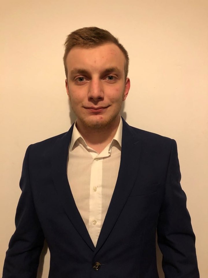 Portret młodego mężczyzny w granatowej marynarce i białej koszuli, stojącego na tle jasnej ściany.