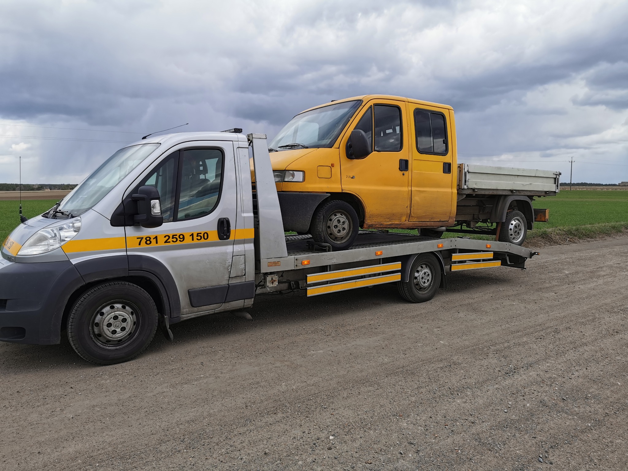 Srebrna laweta z żółtym busem na platformie, zaparkowana na szutrowej drodze, z polami w tle i pochmurnym niebem.