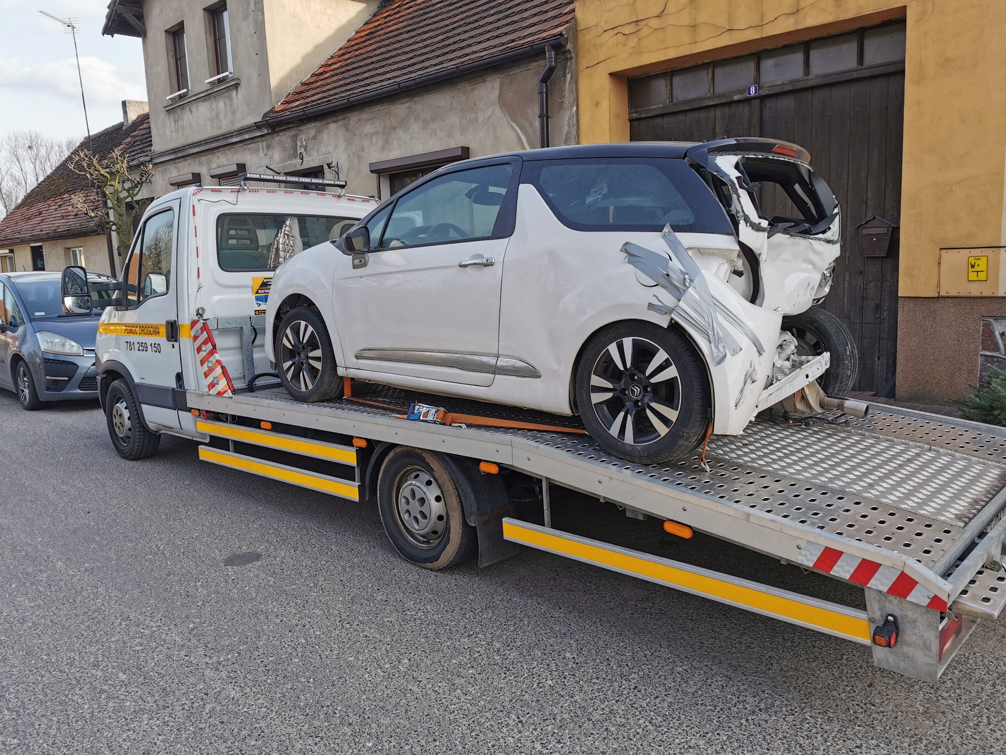 Uszkodzony, biały samochód osobowy z czarnym dachem, zabezpieczony srebrną taśmą, załadowany na lawetę przed budynkiem mieszkalnym.
