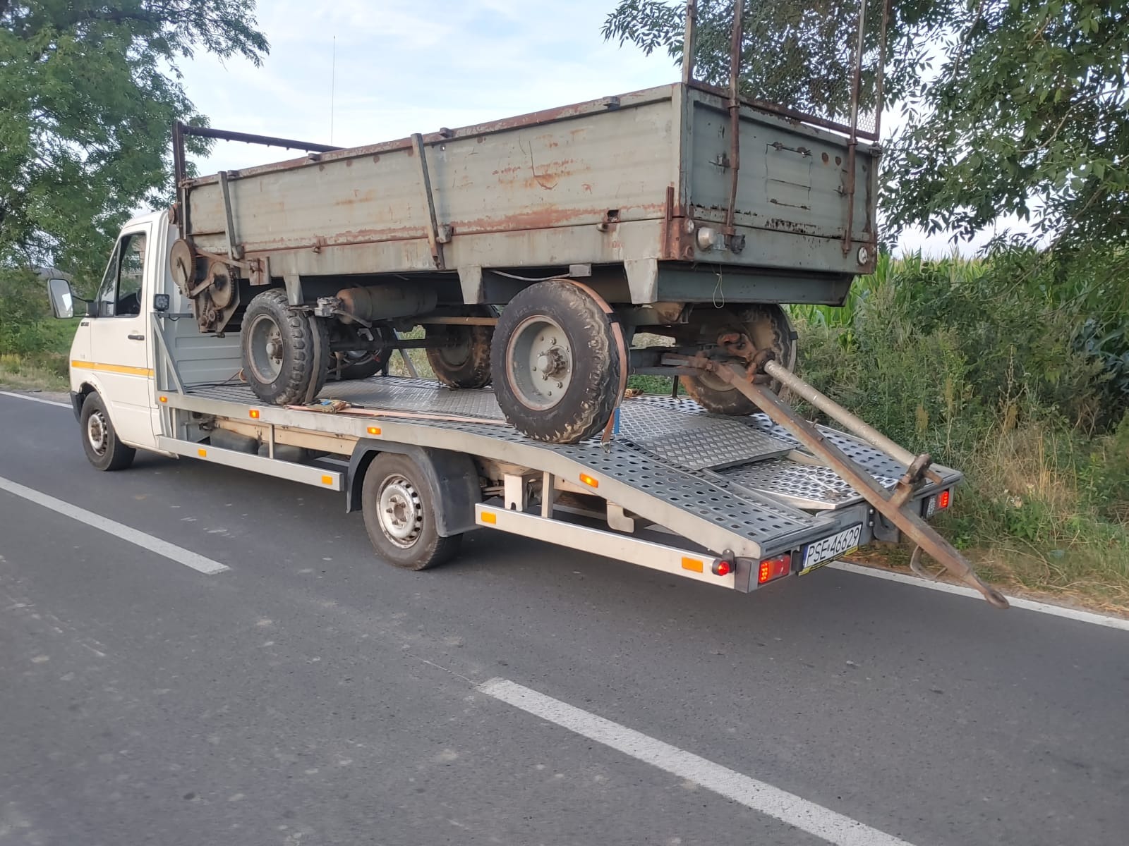 Laweta przewozi stary, skorodowany wóz rolniczy z czterema kołami po asfaltowej drodze, w tle widoczne drzewa i kukurydza.