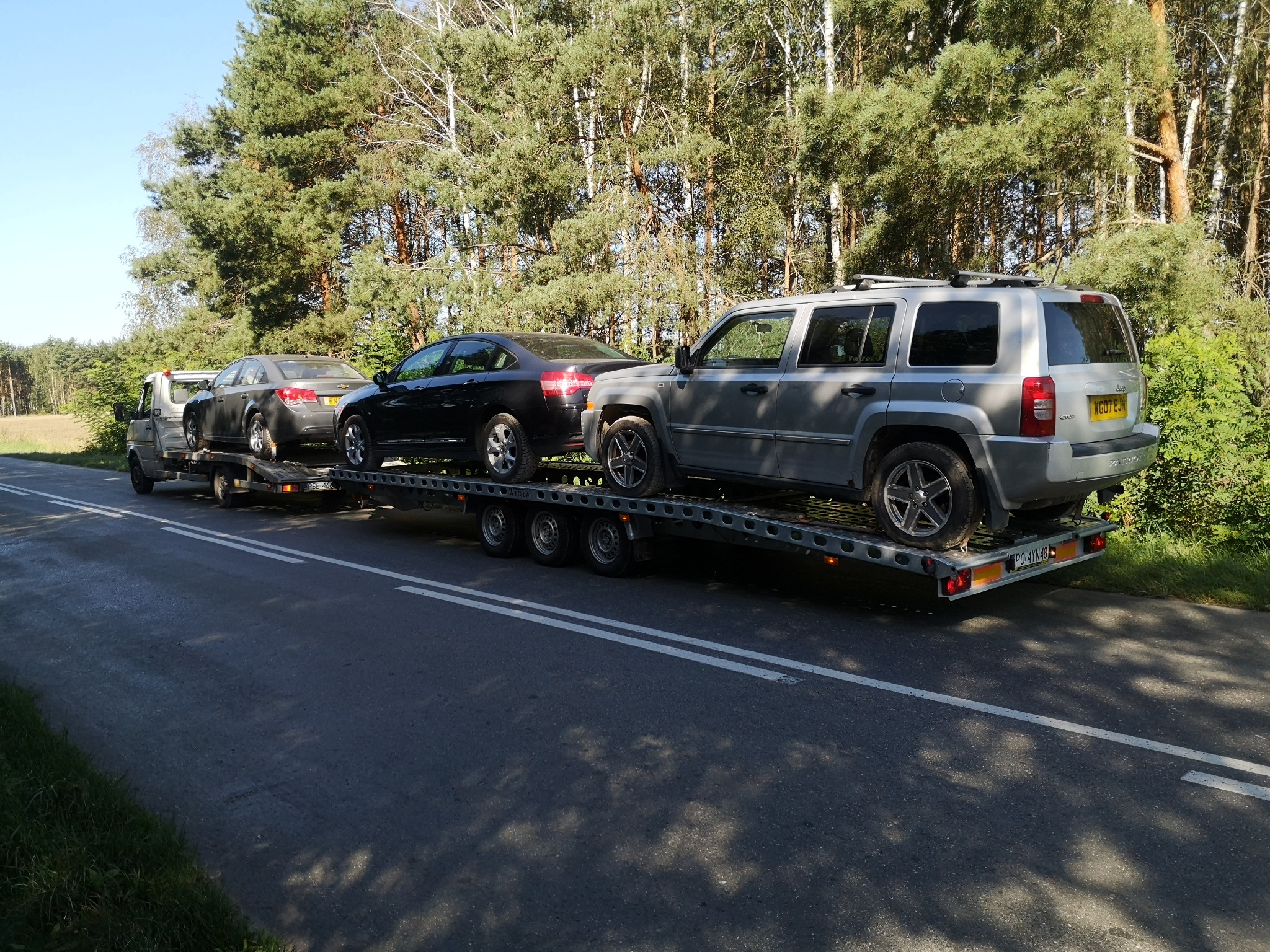 Laweta transportuje trzy samochody osobowe różnej marki i koloru na tle drzew