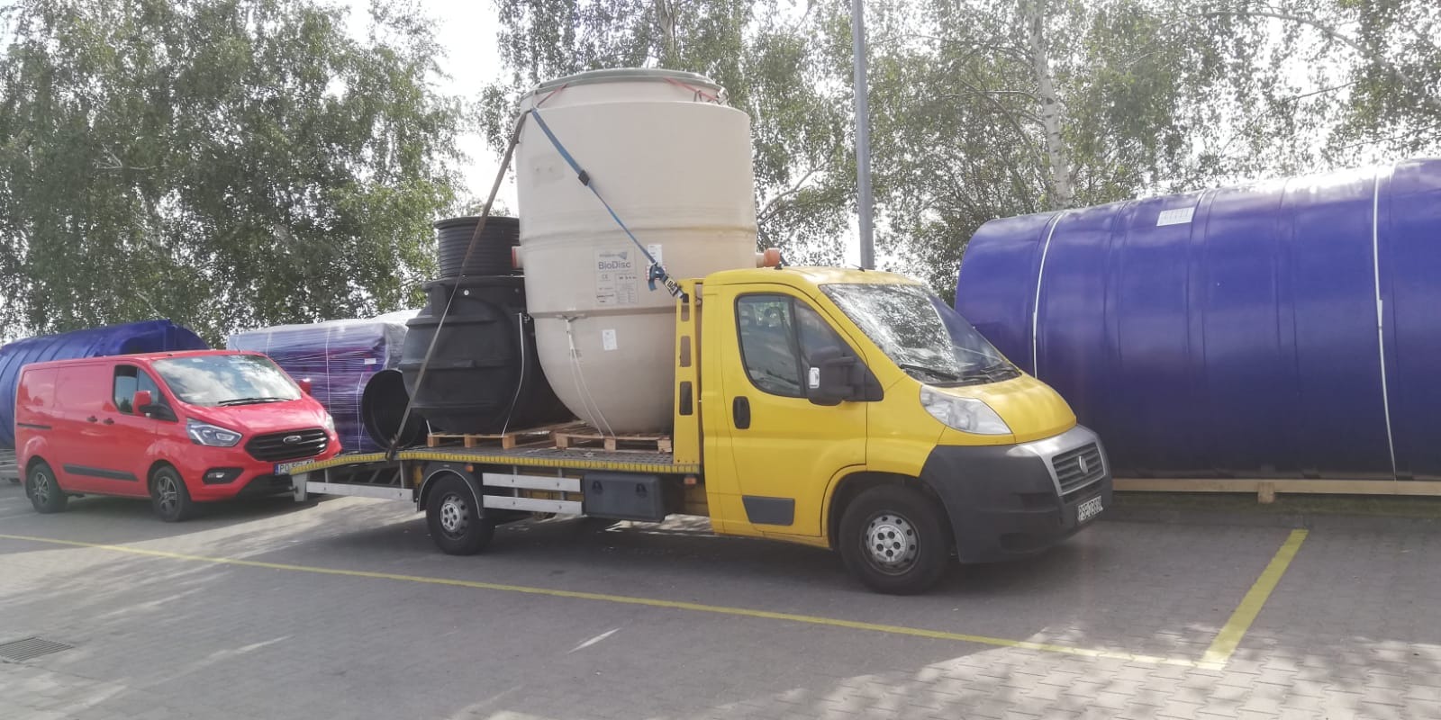 Żółta laweta z załadowanymi zbiornikami na paletach, czerwony bus obok, na parkingu z drzewami w tle.