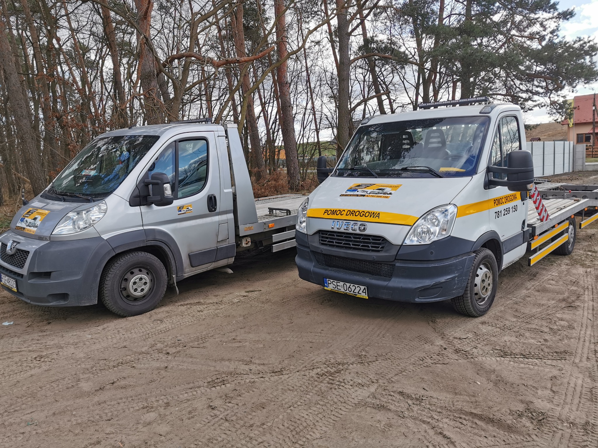 Dwa pojazdy pomocy drogowej, srebrny Peugeot i biały Iveco, stoją obok siebie na piaszczystym terenie z drzewami w tle. Na białym Iveco widoczny napis 'POMOC DROGOWA' i numer telefonu.