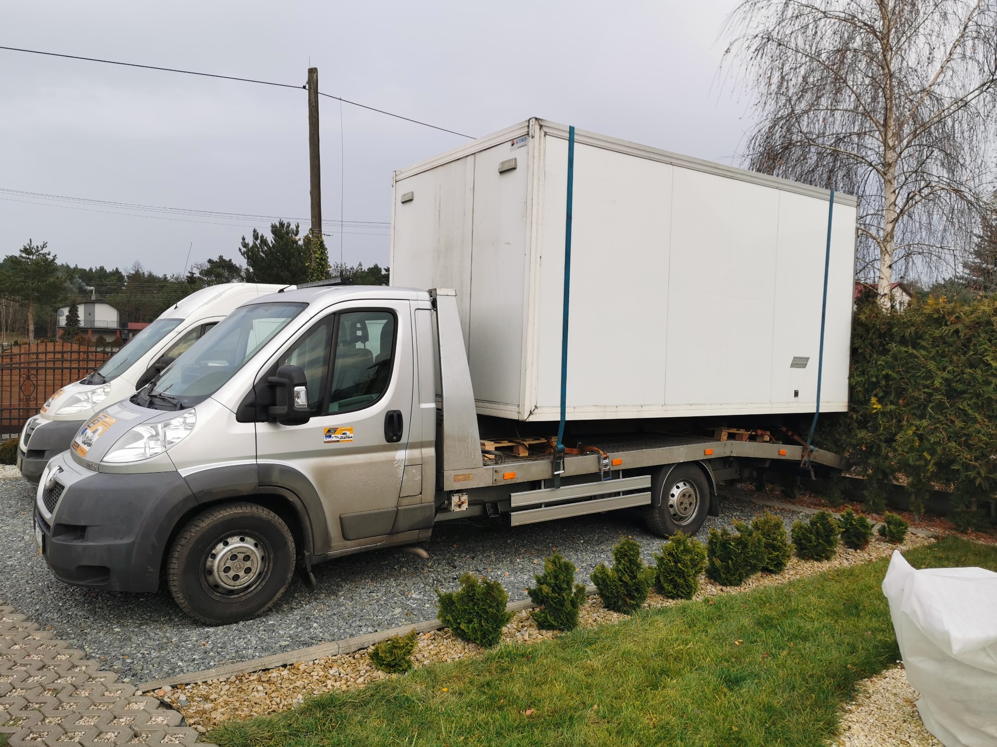 Srebrny samochód dostawczy z białą zabudową kontenerową na platformie, zaparkowany na żwirowym podjeździe, z drugim identycznym pojazdem widocznym w tle.