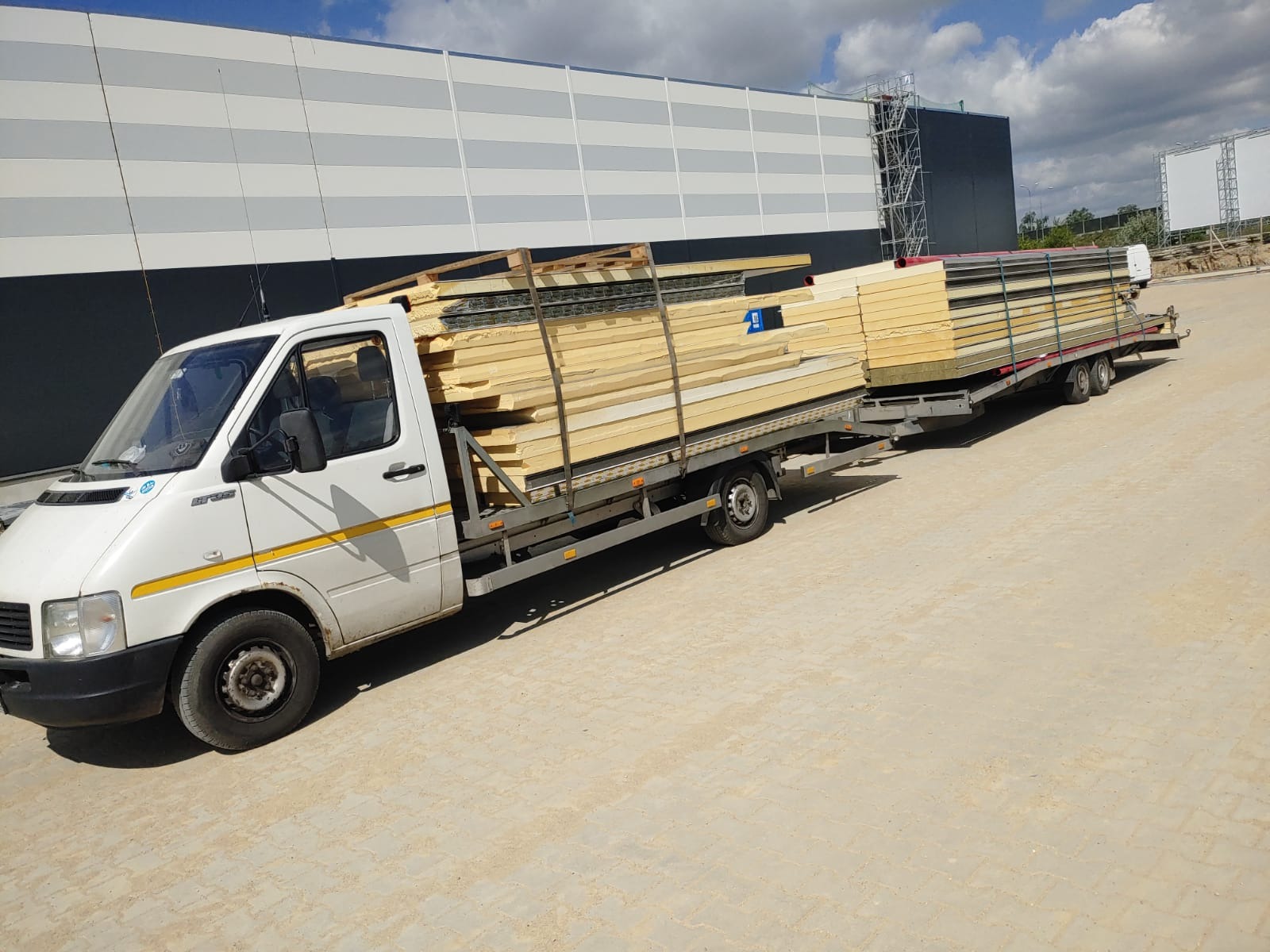 Biała laweta z przyczepą załadowana panelami izolacyjnymi na tle szarego magazynu.