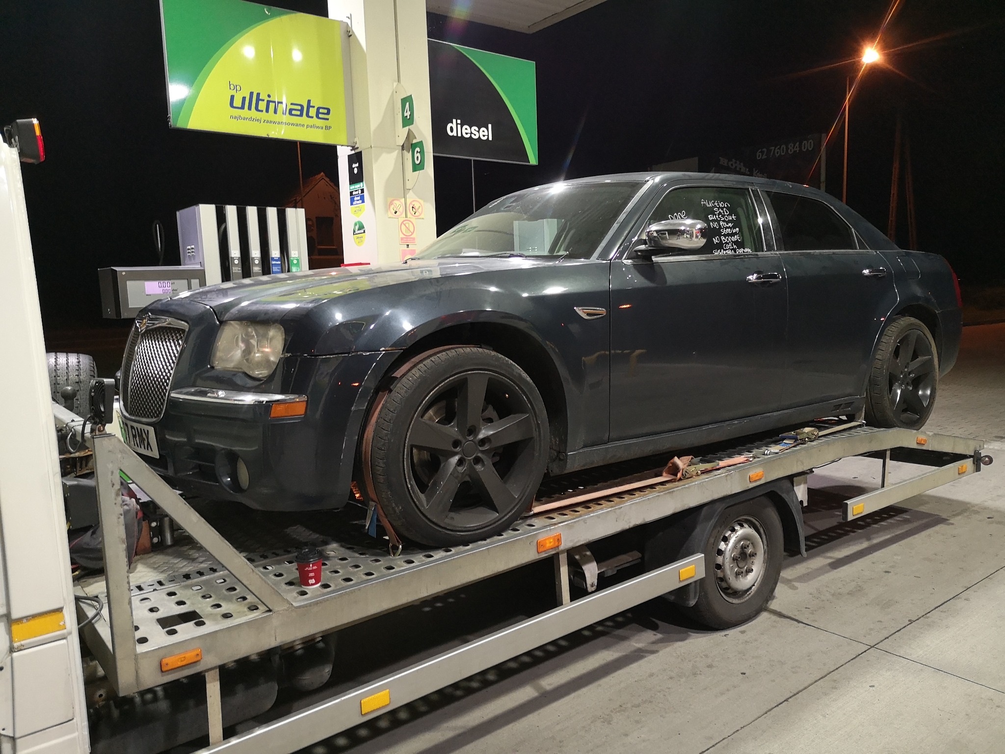 Uszkodzony, ciemnoszary samochód osobowy marki Chrysler, zabezpieczony pasami transportowymi, przewożony na lawecie na stacji paliw BP nocą. Na szybie auta widoczne notatki o uszkodzeniach.