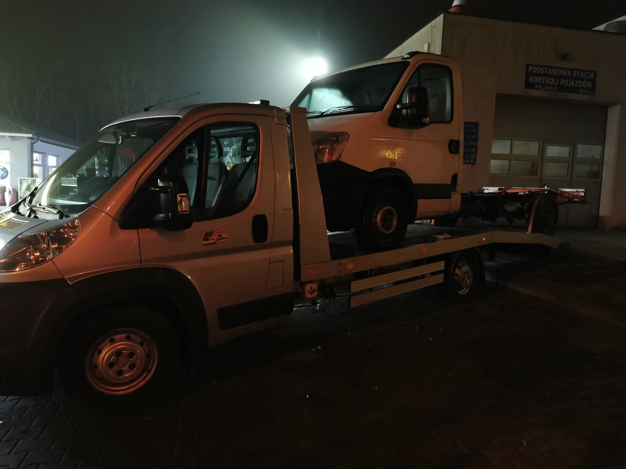 Laweta z białym samochodem dostawczym na platformie, stojąca nocą przed stacją kontroli pojazdów.
