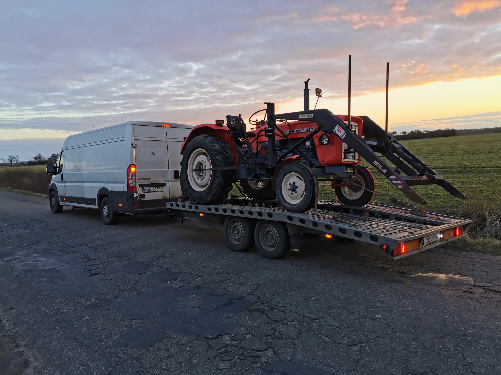 Biały bus z lawetą przewożący czerwony traktor marki Ursus C-360 na tle pola i zachodzącego słońca.