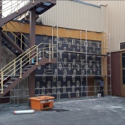 VERSUM GROUP SP. Z O.O. - Prace przygotowawcze do ocieplania elewacji budynku przemysłowego: nałożona warstwa kleju z widocznymi punktami mocowania izolacji, rusztowanie, pracownik przy pracy, pomarańczowy pojemnik...