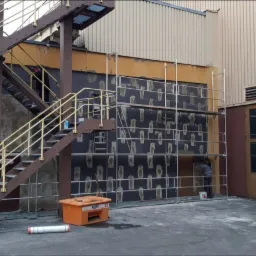 Prace przygotowawcze do ocieplania elewacji budynku przemysłowego: nałożona warstwa kleju z widocznymi punktami mocowania izolacji, rusztowanie, pracownik przy pracy, pomarańczowy pojemnik...