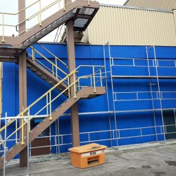 VERSUM GROUP SP. Z O.O. - Niebieska ściana budynku z metalowymi rusztowaniami i schodami z żółtymi poręczami, pomarańczowy pojemnik na pierwszym planie z napisem RUWE 22.