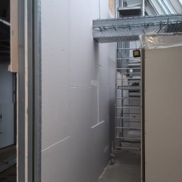 VERSUM GROUP SP. Z O.O. - Montaż płyt gipsowo-kartonowych na metalowym ruszcie w pomieszczeniu z widocznym rusztowaniem i oknem dachowym.