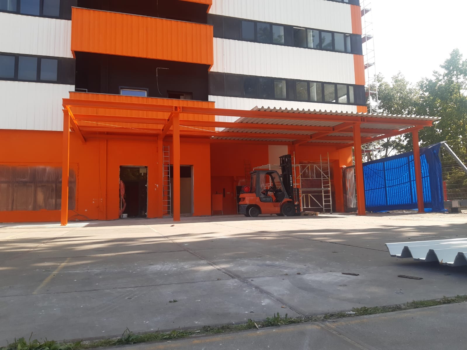 Pomarańczowy budynek biurowy z zadaszeniem, w tle rusztowania i wózek widłowy, widoczne ślady po malowaniu na ścianie.
