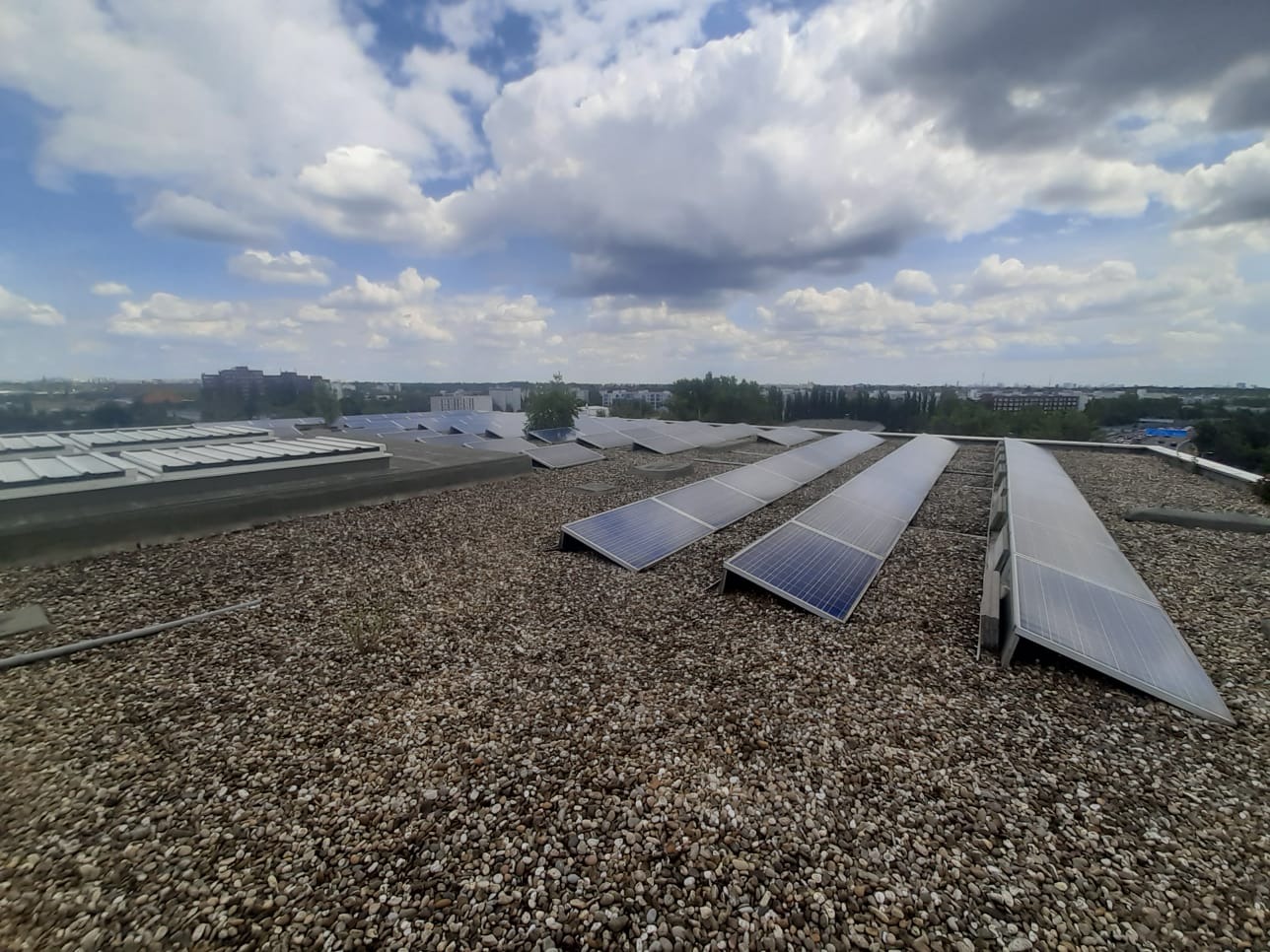 Widok z góry na instalację paneli słonecznych na dachu pokrytym drobnym żwirem, z widoczną panoramą miasta i zachmurzonym niebem.