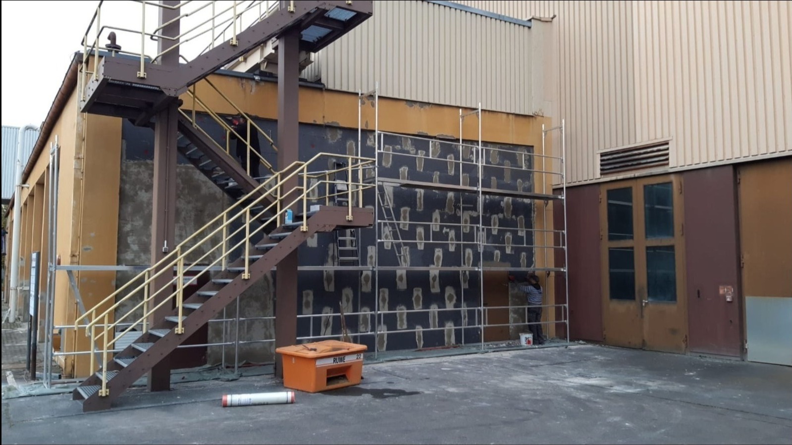 Prace przygotowawcze do ocieplania elewacji budynku przemysłowego: nałożona warstwa kleju z widocznymi punktami mocowania izolacji, rusztowanie, pracownik przy pracy, pomarańczowy pojemnik...