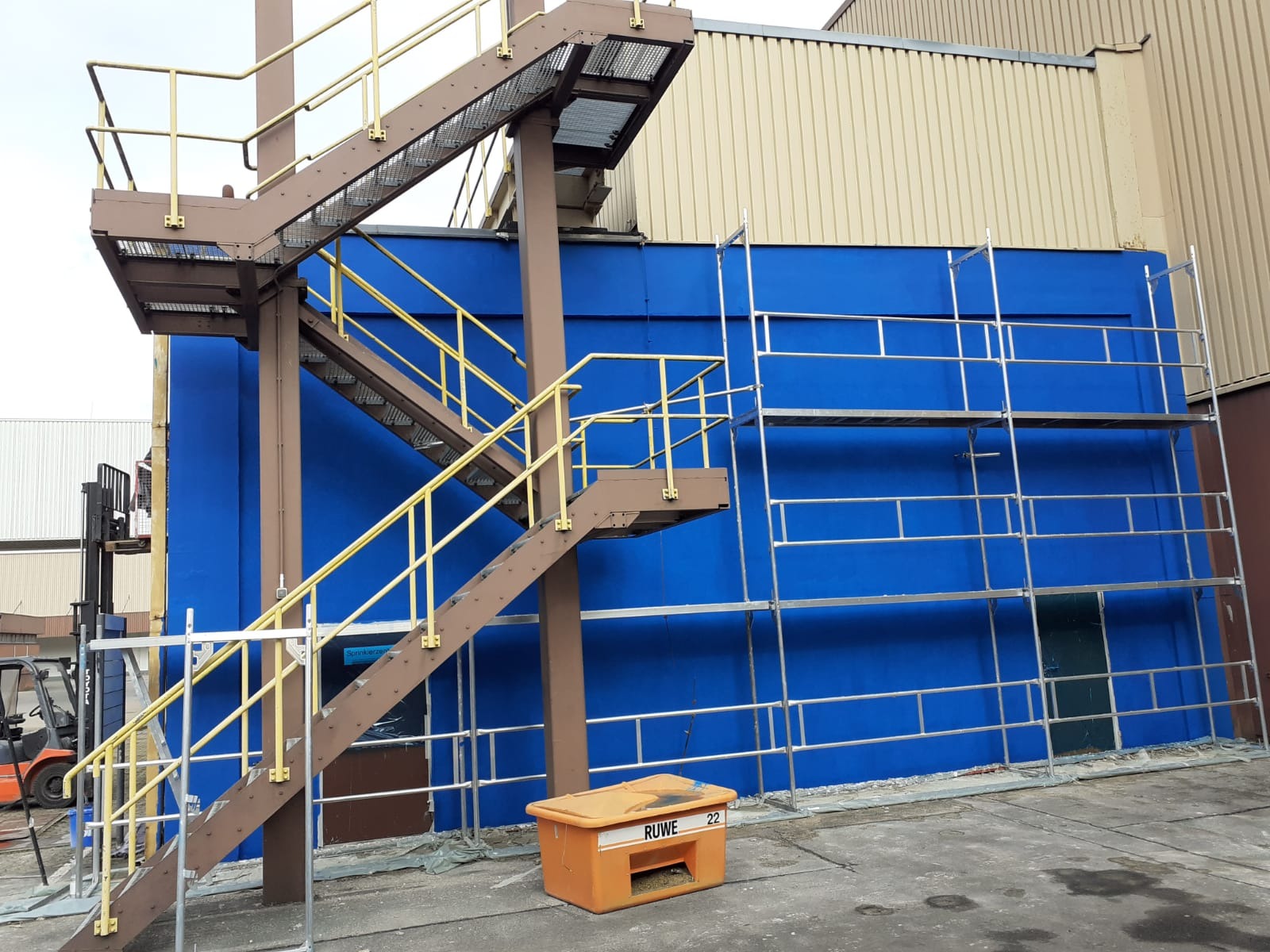 Niebieska ściana budynku z metalowymi rusztowaniami i schodami z żółtymi poręczami, pomarańczowy pojemnik na pierwszym planie z napisem RUWE 22.