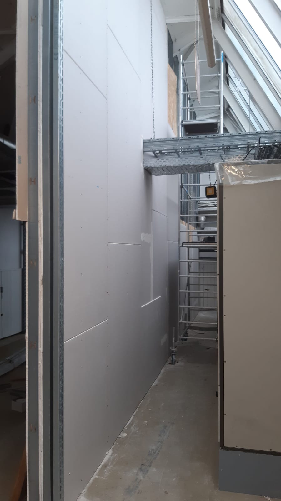 Montaż płyt gipsowo-kartonowych na metalowym ruszcie w pomieszczeniu z widocznym rusztowaniem i oknem dachowym.