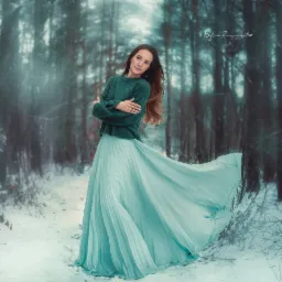 Kobieta w zielonym swetrze i turkusowej, plisowanej sp&oacute;dnicy pozuje w zimowym lesie, śnieżny krajobraz, subtelne oświetlenie, widoczne logo fotografa w prawym g&oacute;rnym rogu.