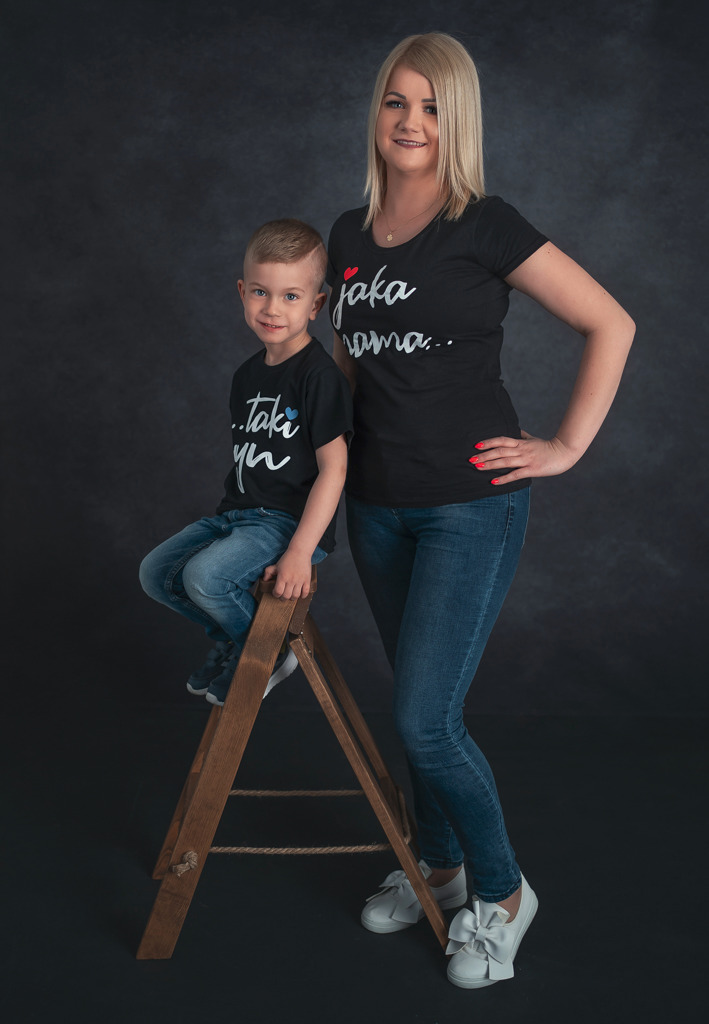 Portret matki z synem w dopasowanych czarnych koszulkach z napisami, chłopiec siedzi na drewnianej drabince na tle ciemnego, neutralnego tła.