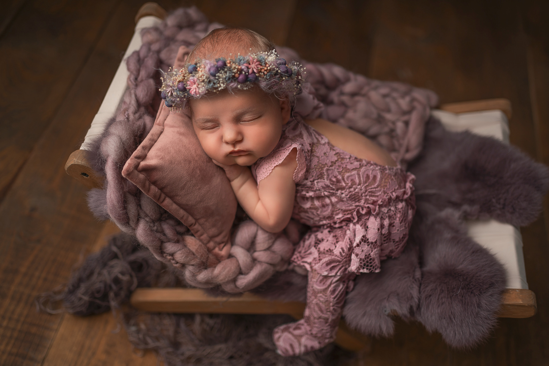 Śpiąca noworodka w koronie z kwiatów, ubrana w koronkowy kombinezon, ułożona na małym łóżeczku z poduszką i miękkimi kocykami w odcieniach fioletu i różu.