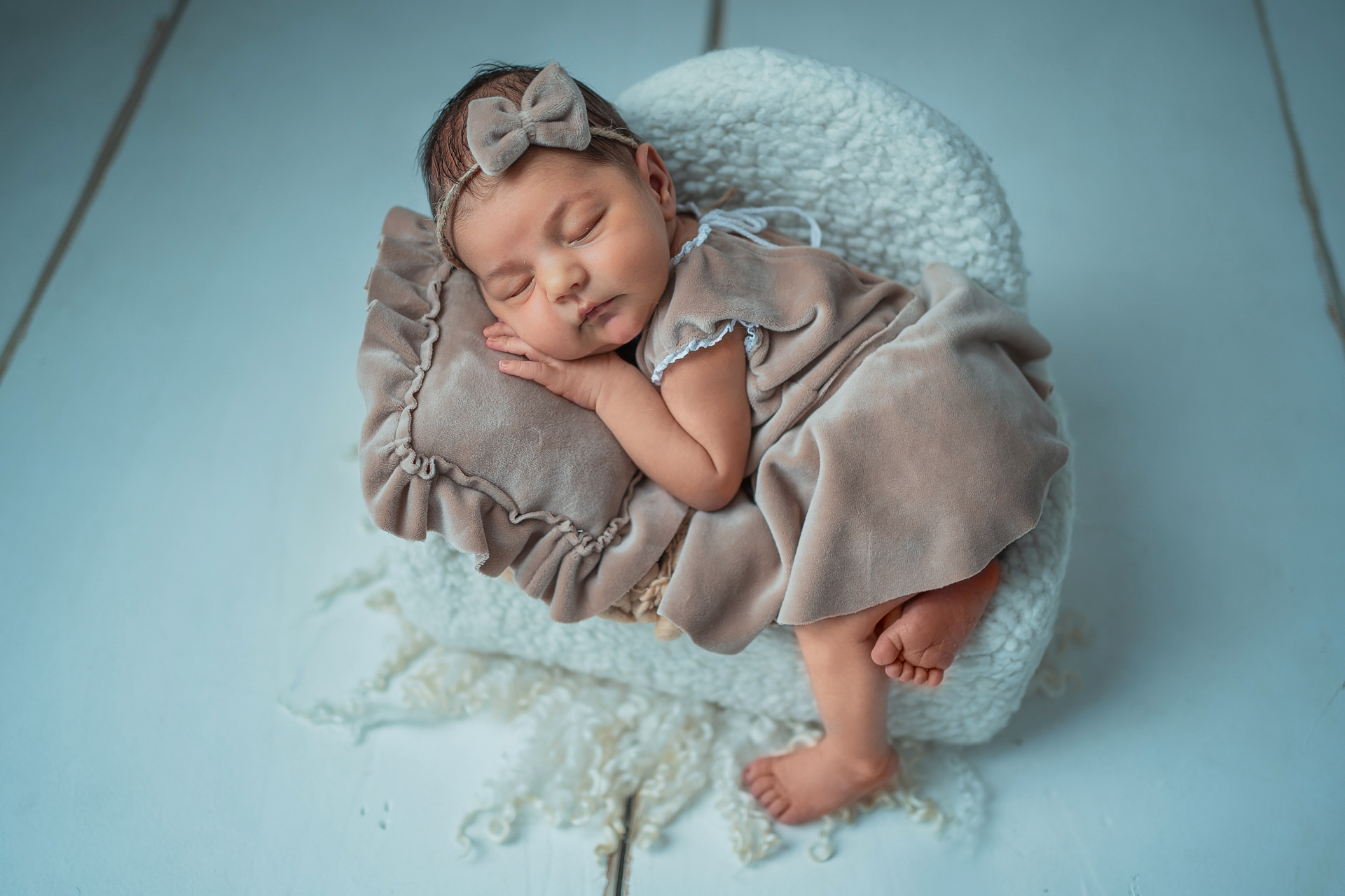Śpiąca nowo narodzona dziewczynka w beżowej sukience i z kokardą na głowie, ułożona na puszystym, białym kocyku i poduszce z falbankami.