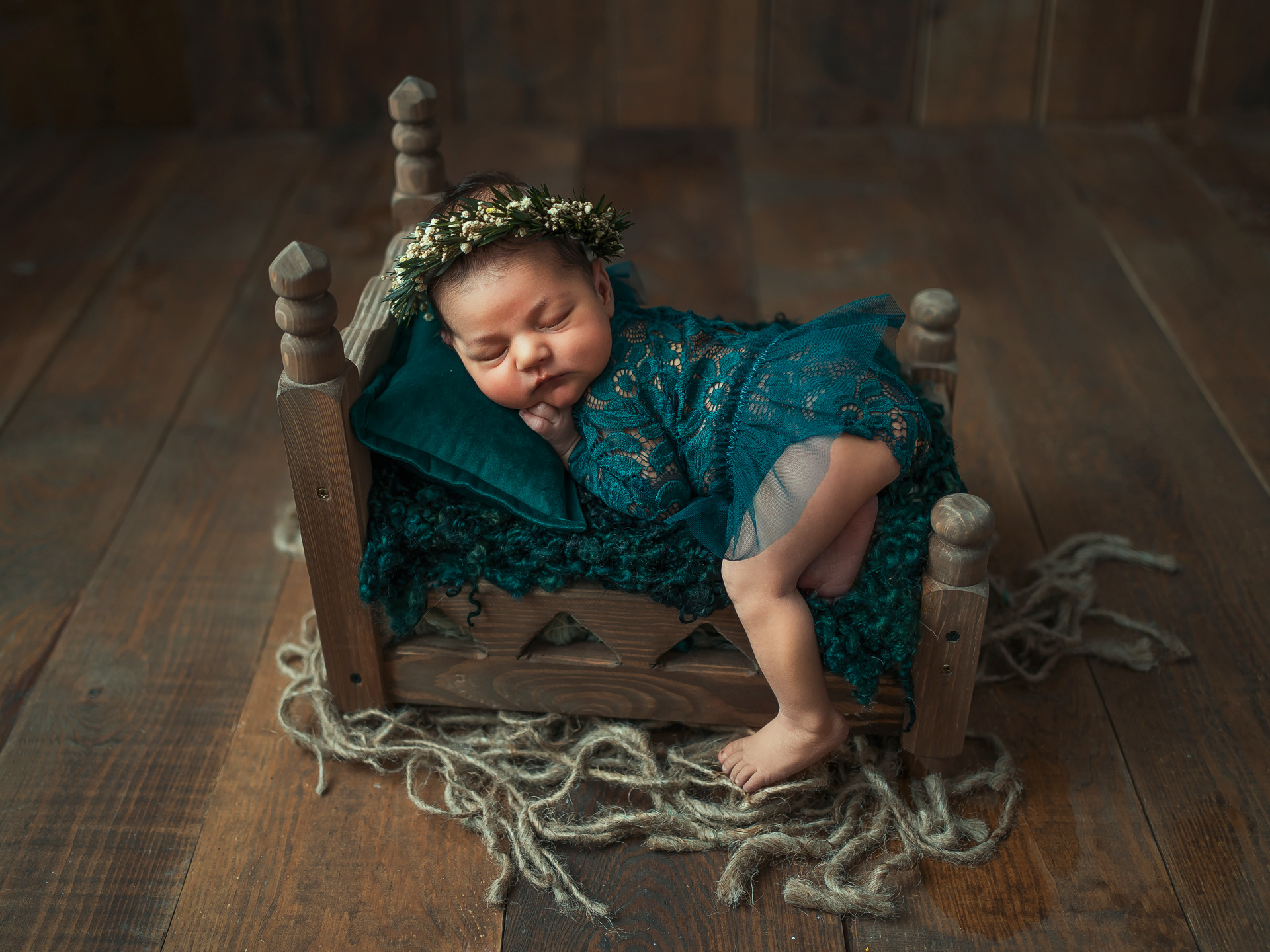 Noworodek w koronie z kwiatów śpiący na małym drewnianym łóżku z zieloną poduszką i pledem, na drewnianej podłodze z jutowymi sznurami.