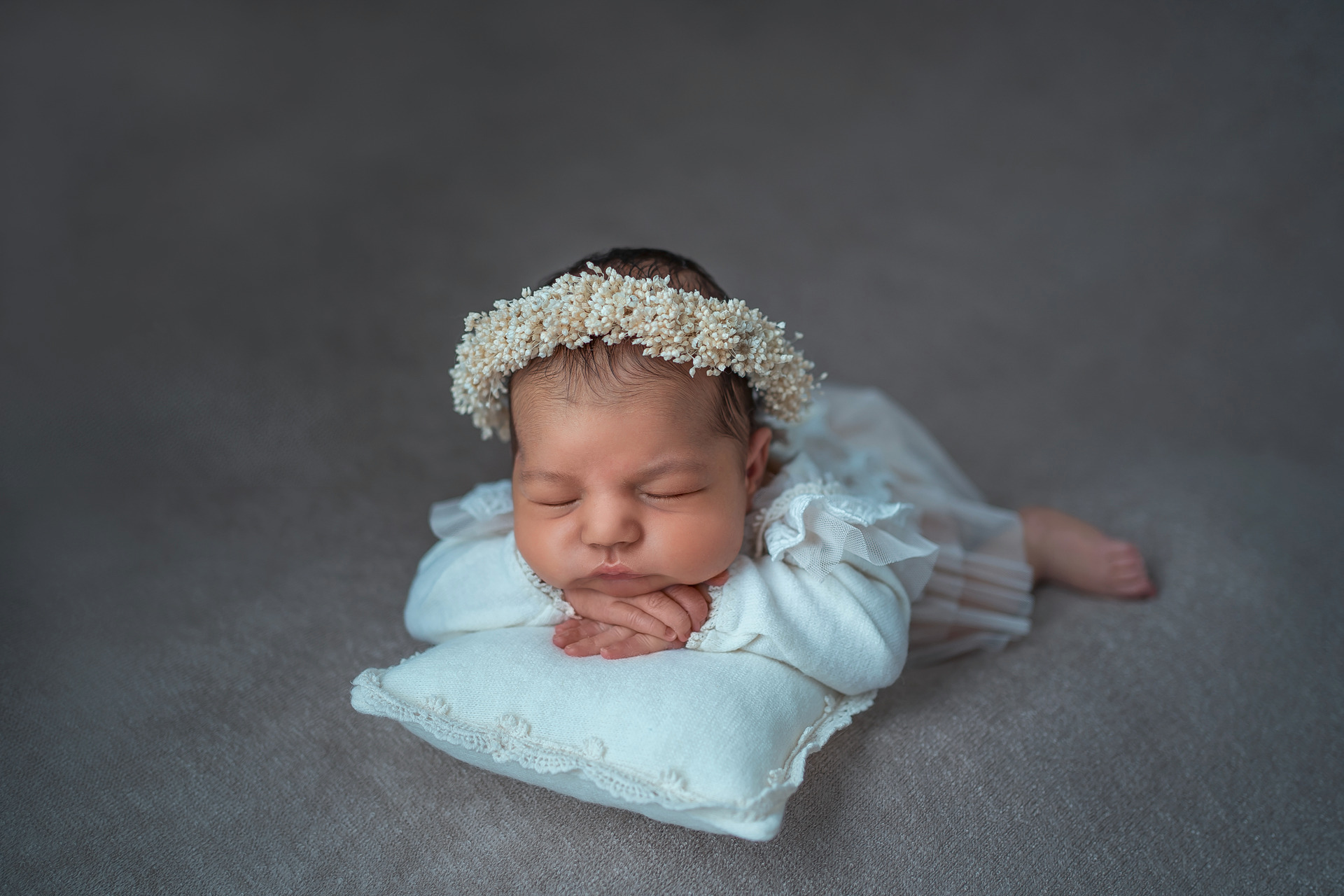 Śpiący noworodek w białej sukience z koronkowymi falbankami i wiankiem z drobnych kwiatów na głowie, ułożony na małej, kwadratowej poduszce z koronkowym brzegiem, na szarym tle.