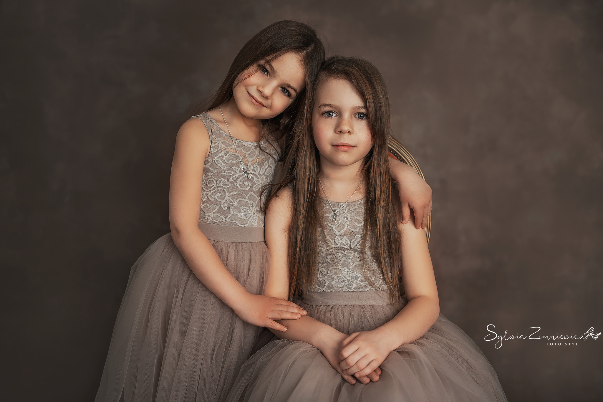 Portret dwóch dziewczynek w tiulowych sukienkach z koronkową górą, obejmujących się na tle ciemnego, teksturowanego tła. Jedna z dziewczynek opiera głowę o ramię drugiej.