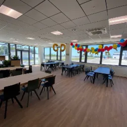 Jasne, przestronne biuro z drewnianą podłogą, dużymi oknami, stołami i krzesłami, udekorowane balonami z okazji otwarcia lub jubileuszu.