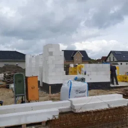 Wznoszenie ścian domu z białych bloczków, widoczne elementy zbrojenia, paleta z bloczkami, worki z zaprawą, oraz fragment elewacji z ciemnej cegły klinkierowej.