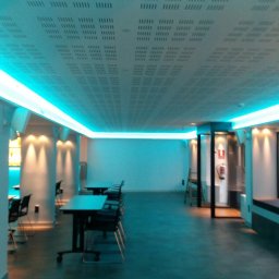 Jadański Bartlomiej - Sala z sufitem z płyty karton gips z perforacją i oświetleniem LED w kolorze turkusowym, widoczne stoły i krzesła.