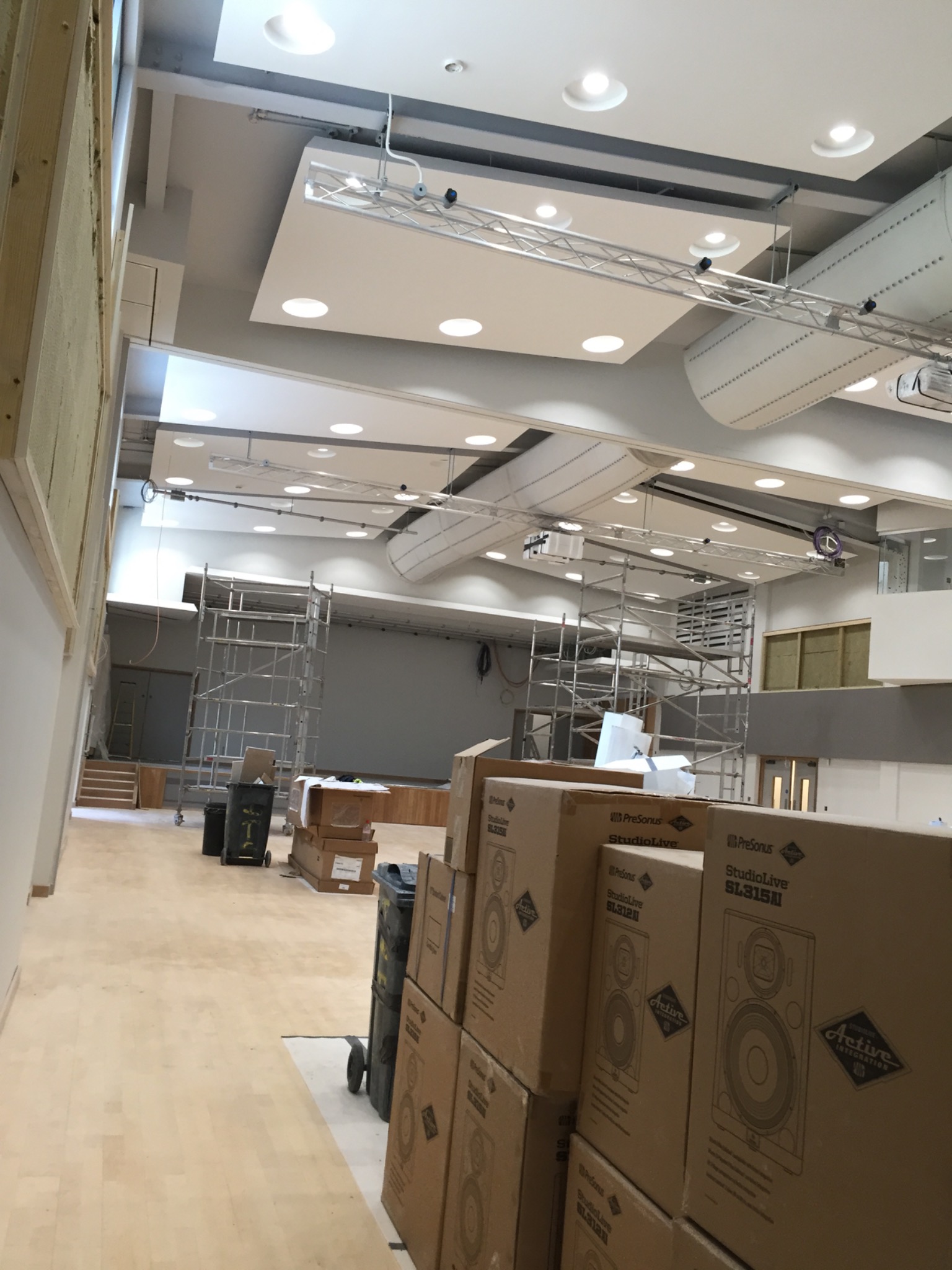 Wnętrze sali widowiskowej w trakcie prac wykończeniowych: widoczne rusztowania, elementy konstrukcji sufitu z oświetleniem punktowym oraz kartony z głośnikami, częściowo zasłaniające widok.