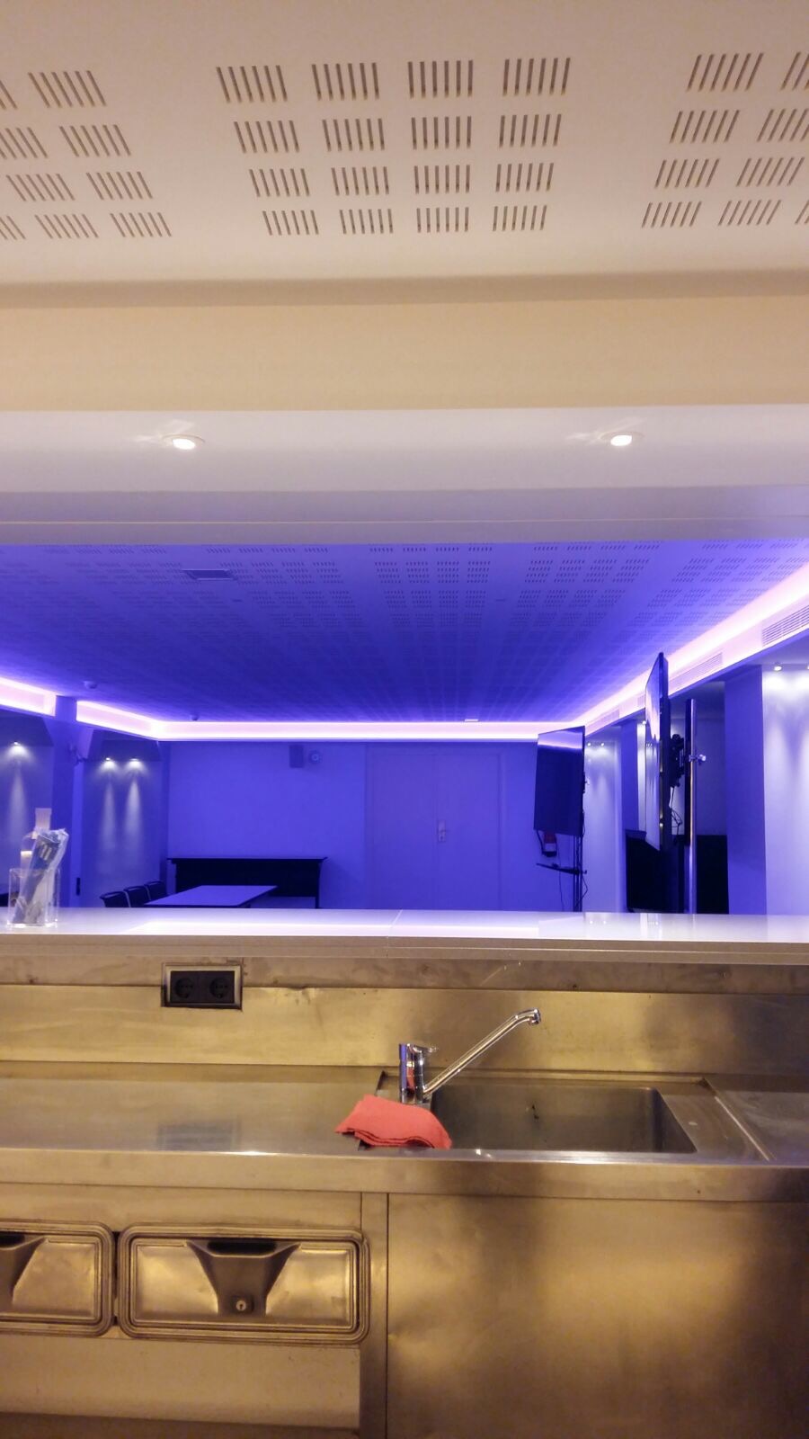 Metalowa umywalka i blat kuchenny widziane od dołu, w tle pomieszczenie z podwieszanym sufitem oświetlonym niebieskim światłem LED i perforowaną strukturą.