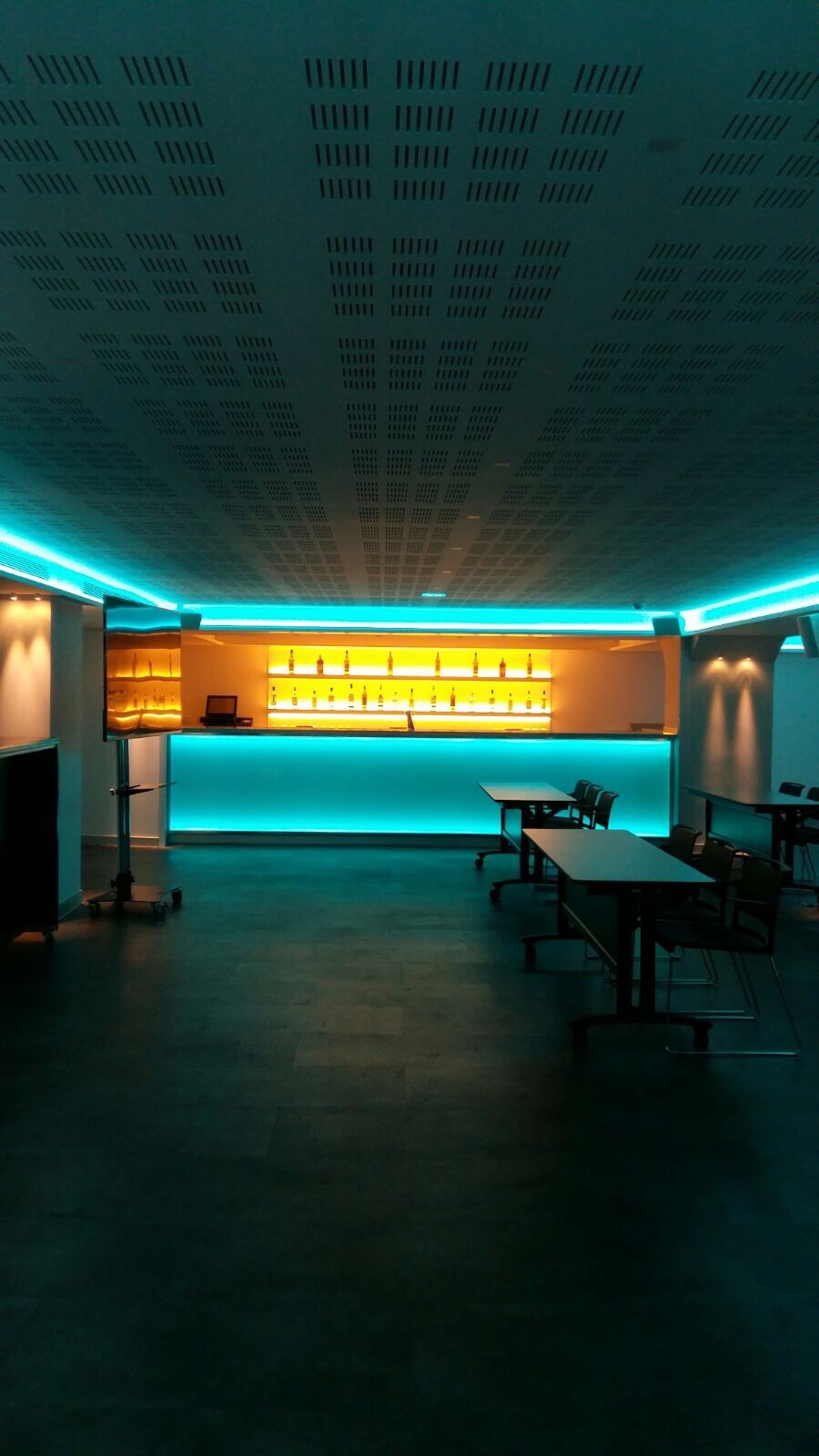 Wnętrze lokalu z barem podświetlonym turkusowym światłem LED, perforowany sufit z oświetleniem, ciemna podłoga i kilka stolików.