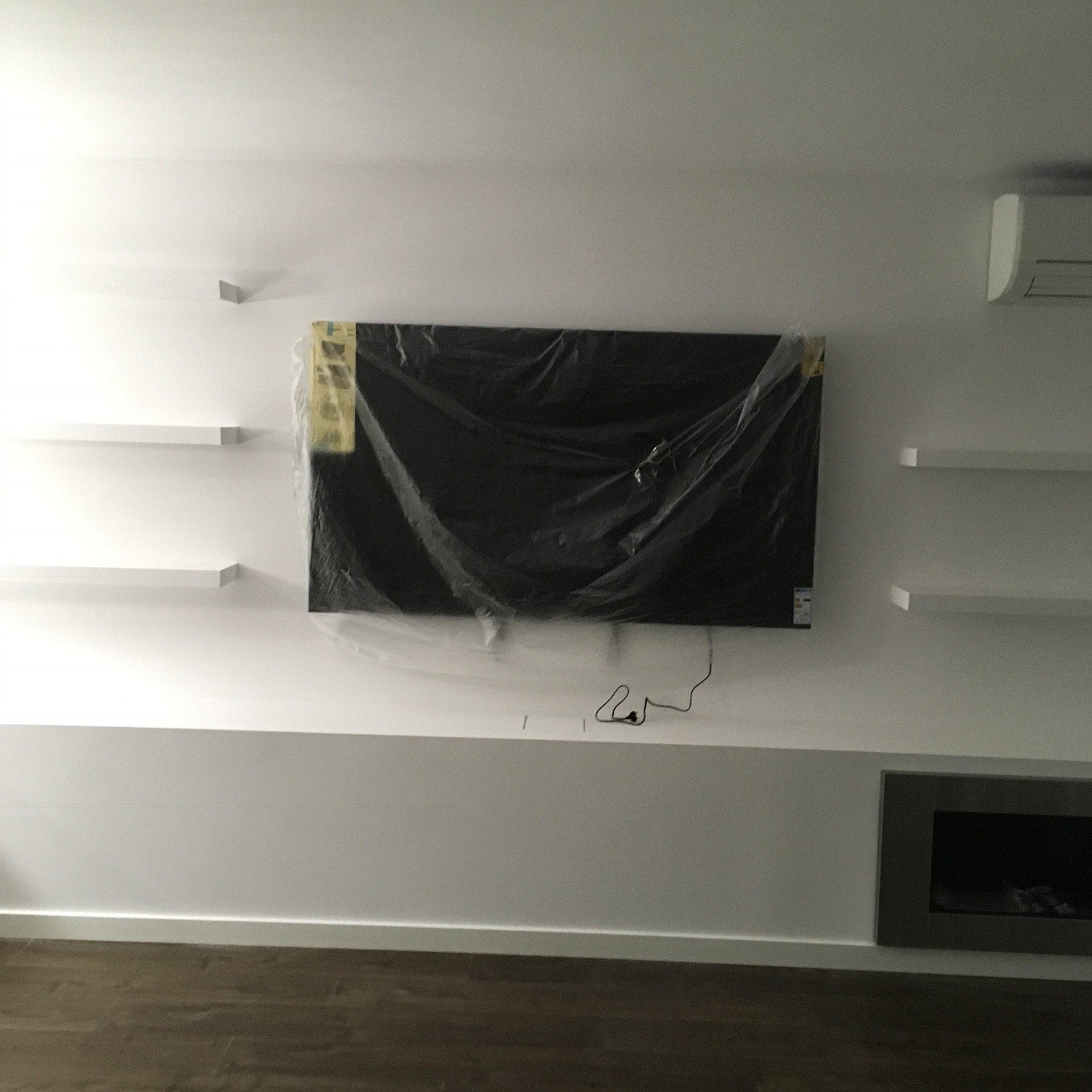 Biała ściana z wnęką na kominek, telewizorem owiniętym folią ochronną i minimalistycznymi półkami.