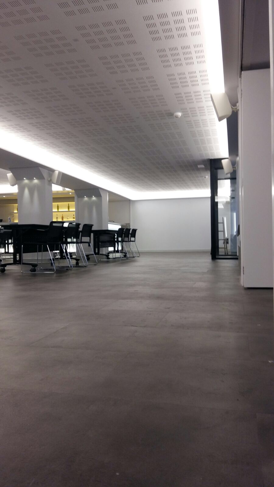 Wnętrze biurowe z podwieszanym sufitem kasetonowym oświetlonym liniowymi lampami LED, widoczne stoły z krzesłami na kółkach i filary.