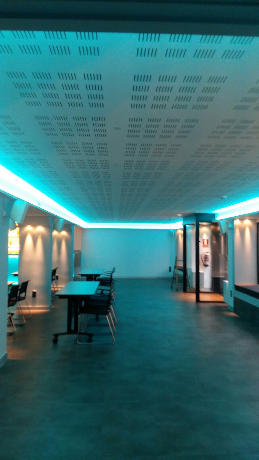 Sala z sufitem z płyty karton gips z perforacją i oświetleniem LED w kolorze turkusowym, widoczne stoły i krzesła.