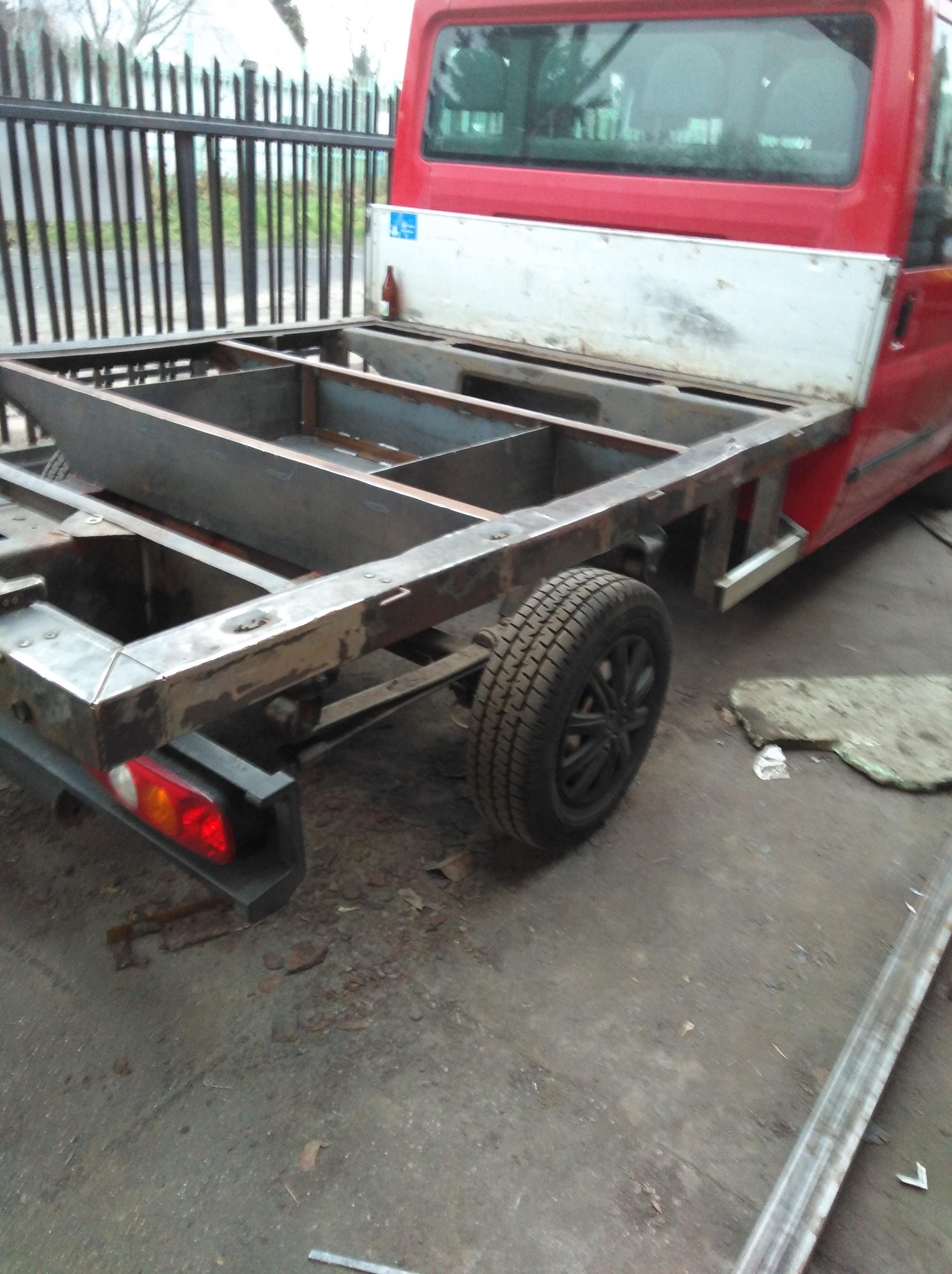 Metalowa konstrukcja ramy pojazdu na podwoziu czerwonego dostawczaka, widoczne ślady spawania i obróbki metalu, w tle ogrodzenie posesji.