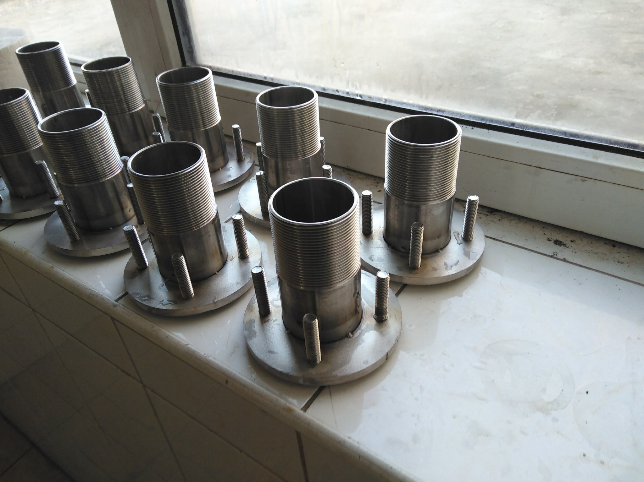 Szereg identycznych, metalowych elementów z gwintowanym cylindrem, okrągłą podstawą i trzema śrubami, ułożonych na białej, kafelkowanej powierzchni przy oknie.