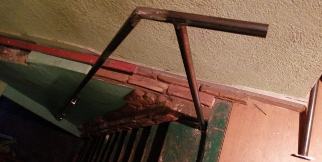 Wykadrowany widok fragmentu konstrukcji metalowej na schodach, surowy spaw na łączeniach rur, nieestetyczne otoczenie z cegłą i odrapaną farbą.